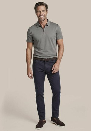 Mann stehend, lächelnd, trägt ein graues kurzärmeliges Polo, dunkelblaue Hose mit braunem Gürtel und dunkelbraune Slipper vor neutralem Hintergrund.