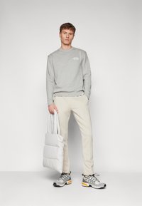 Felpa grigia, pantaloni beige, porta una borsa imbottita grigia chiaro e indossa sneakers grigie con accenti arancioni. Design semplice e minimalista.