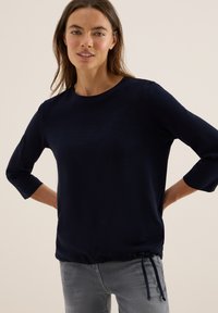 Kvinna i en marinblå långärmad tröja med dragsko och ljusgrå jeans, stående med händerna bakom ryggen mot en beige bakgrund.