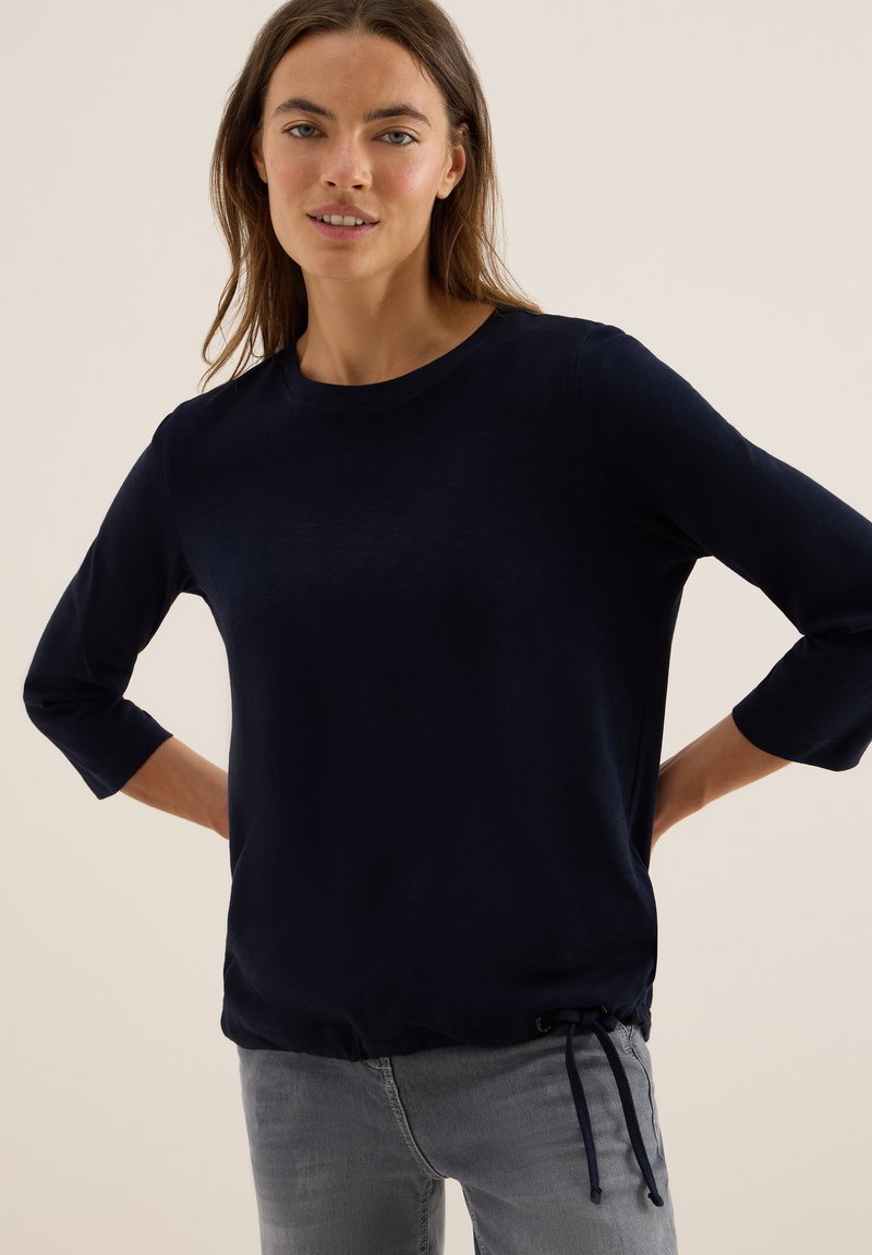 Kvinna i en marinblå långärmad tröja med dragsko och ljusgrå jeans, stående med händerna bakom ryggen mot en beige bakgrund.