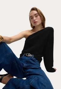 Un pull en tricot noir avec un col asymétrique, associé à un jean en denim bleu ample et une ceinture noire avec une boucle en or.