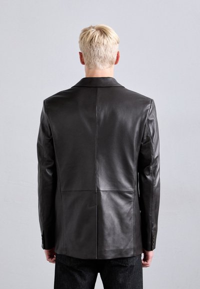 Man with short blonde hair wearing a fitted black leather jacket, shown from the back against a plain gray background.