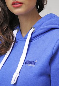 Sudadera azul con una textura suave, cordones blancos y el logotipo bordado de "SuperDry" en azul oscuro. Presenta detalles metálicos cerca de la capucha.