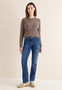 Un pull beige en maille côtelée associé à un jean droit bleu, avec un détail de lien à l'avant et un lavage légèrement décoloré. Des baskets beiges complètent la tenue.
