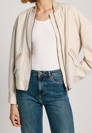 Femme portant une veste crème zippée sur un débardeur blanc et un jean bleu taille haute, main posée sur la hanche, fond neutre.