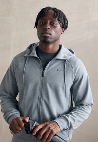 Gray zip-up hoodie with a hood, made of soft material. Features a small logo on the chest and a relaxed fit. User holds a water bottle.