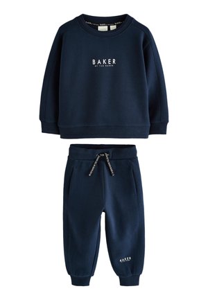 Ung dreng smilende, stående med den ene hånd i lommen, iført marineblå sweatshirt og bukser med hvide sneakers mod en ensfarvet baggrund.