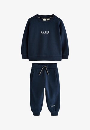 Ung dreng smilende, stående med den ene hånd i lommen, iført marineblå sweatshirt og bukser med hvide sneakers mod en ensfarvet baggrund.