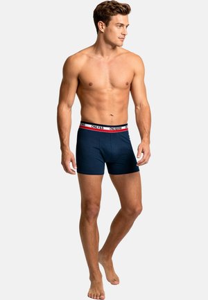 Ung mand med muskuløs krop iført navyblå boxershorts med en hvid, rød og sort logoelastik, stående barfodet på en ensfarvet baggrund.