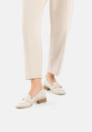Beige leather loafers with a rounded toe, low stacked heel, and decorative strap. Worn with light-colored loose-fitting trousers.