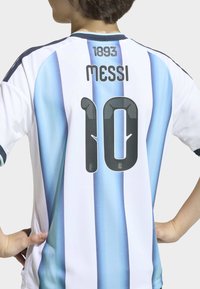 Młoda osoba w koszulce piłkarskiej Argentyny z niebieskimi i białymi pionowymi paskami, z nazwiskiem "Messi", numerem 10 oraz tekstem "1893" na plecach.