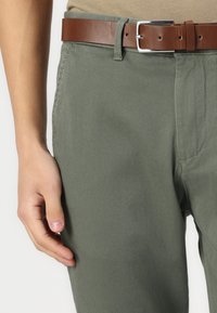 Pantalon texturé vert olive avec une finition lisse, mettant en avant une ceinture en cuir marron avec une boucle en argent. Une main repose légèrement sur le tissu.