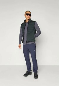 Ανθρωπος φορώντας σκούρα γυαλιά ηλίου, μαύρο γιλέκο, γκρι μακρυμάνικο μπλουζάκι, μπλε φόρμες και μαύρα αθλητικά παπούτσια, στέκεται μπροστά σε απλό φόντο.