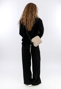 Borsa a tracolla beige trapuntata con superficie testurizzata e tracolla nera regolabile, indossata su un outfit nero morbido, composto da pantaloni larghi e una maglia.
