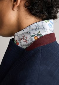 Veste tailleur bleu marine avec un col bordeaux. Col intérieur blanc orné de motifs floraux en vert, rouge et bleu. Tissu texturé.
