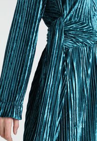 Tyrkysové sametové šaty s vertikálními sklady, dlouhými rukávy a nařasenými detaily v pase. Mají strukturovaný lesk a pružné manžety.