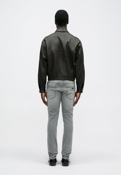 Σκούρα δερμάτινη τζάκετ με ελαστική μέση, γκρι τζιν παντελόνι και μαύρα παπούτσια, εμφανιζόμενα από πίσω σε ανοιχτό φόντο.