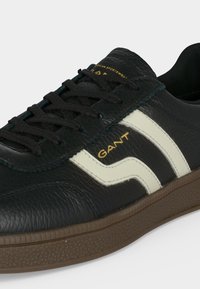 Baskets en cuir noir avec des bandes blanches sur les côtés, semelle marron texturée, lacets noirs et logo doré "GANT" sur le côté.