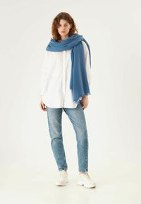 Écharpe bleue en tissu doux, portée sur une chemise blanche à boutons, associée à un jean bleu clair et des baskets blanches, sur un fond neutre.