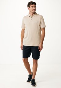 Camisa polo beige con cuello y tres botones, combinada con pantalones cortos negros. El modelo lleva zapatillas negras, mostrando un atuendo casual.