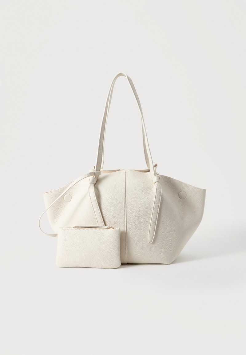 Sac cabas en cuir texturé couleur crème avec deux longues poignées et une petite pochette zippée assortie à l'avant, sur un fond uni.
