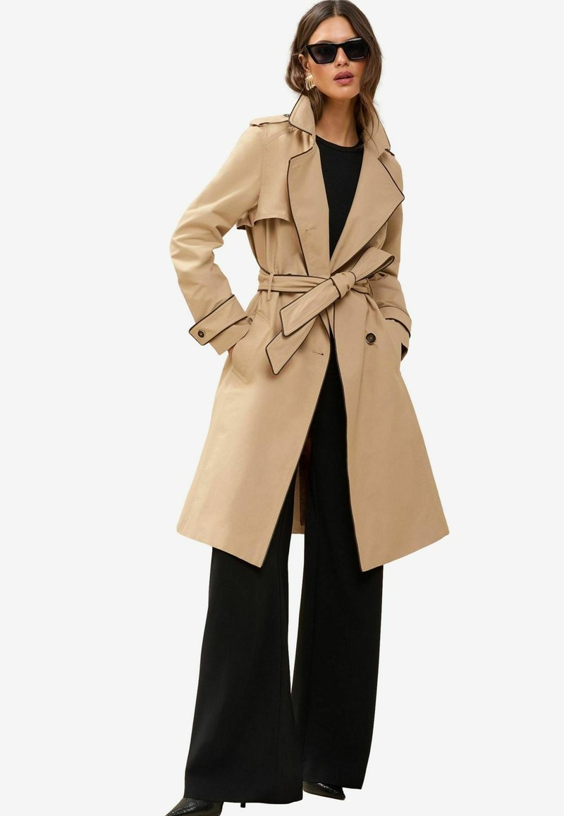 Beige trench kabát fekete kiegészítőkkel, dupla mellényes dizájn, széles hajtókák és levehető öv, fekete, széles lábú nadrággal párosítva.