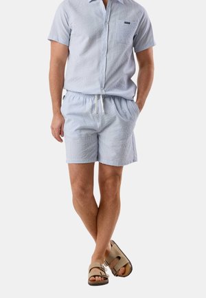 Mann trägt ein hellblaues, strukturiertes kurzärmliges Hemd mit Knöpfen, dazu passende Shorts und beige Sandalen mit zwei Riemen, Hände in den Taschen.