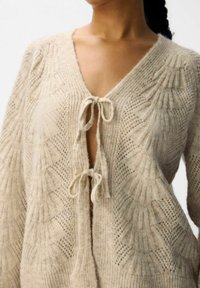 Gebreide cardigan in lichtbeige, met een V-hals, voorste strik en textuurpatronen met opengehaakte details op de mouwen en het lichaam.