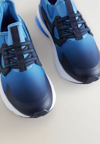 Baskets de sport bleues avec un design dégradé, présentant une texture lisse, des lacets texturés et un capuchon en caoutchouc bleu marine foncé ainsi que des accents.