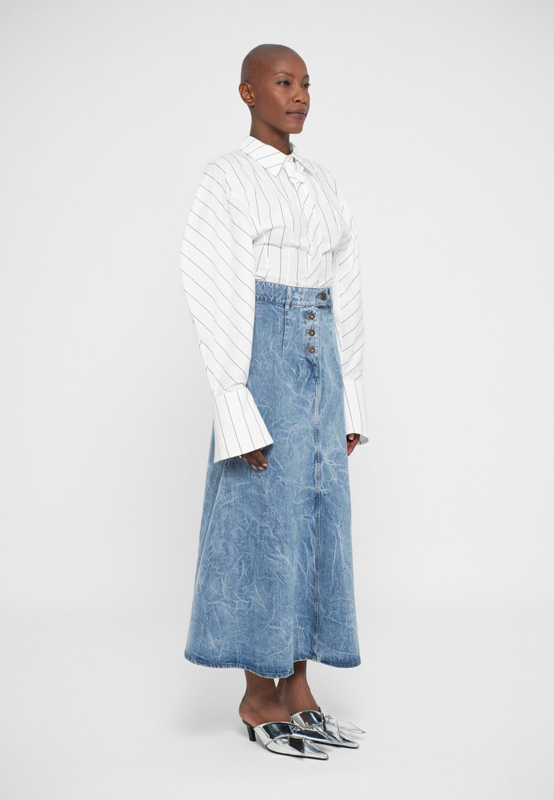 Kvinna med rakad huvud, iklädd en vit randig blus med överdimensionerade manschetter, högmidjad knäppt blå jeanskjol och silverfärgade spetsiga klackar, stående.