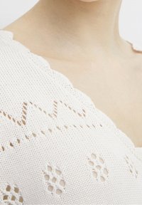 Light beige knitted top featuring a scalloped neckline and openwork patterns, including geometric shapes and cutouts.