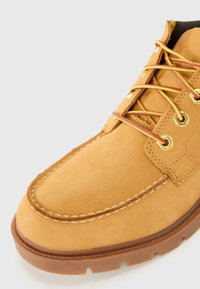 Tan suède laar met ronde neus, contrasterende witte stiksels en metalen ösen. Veters zijn geel met rode accenten. Rubberen zool met grip.
