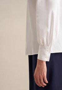 Blusa bianca con maniche lunghe a sbuffo, polsini con bottone e texture liscia. La blusa è abbinata a una gonna blu navy, che presenta un design pulito e minimalista.