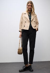 Donna in piedi contro un muro bianco, indossa un giubbino corto beige, una maglietta verde scura, pantaloni neri e scarpe nere, mentre tiene una borsa a spalla in suede beige.
