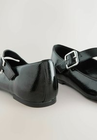 Fekete lakkbőr lapos cipő lekerekített orral, állítható pánttal és alacsony sarokkal. Fényes felülettel és ezüst csattal rendelkezik.