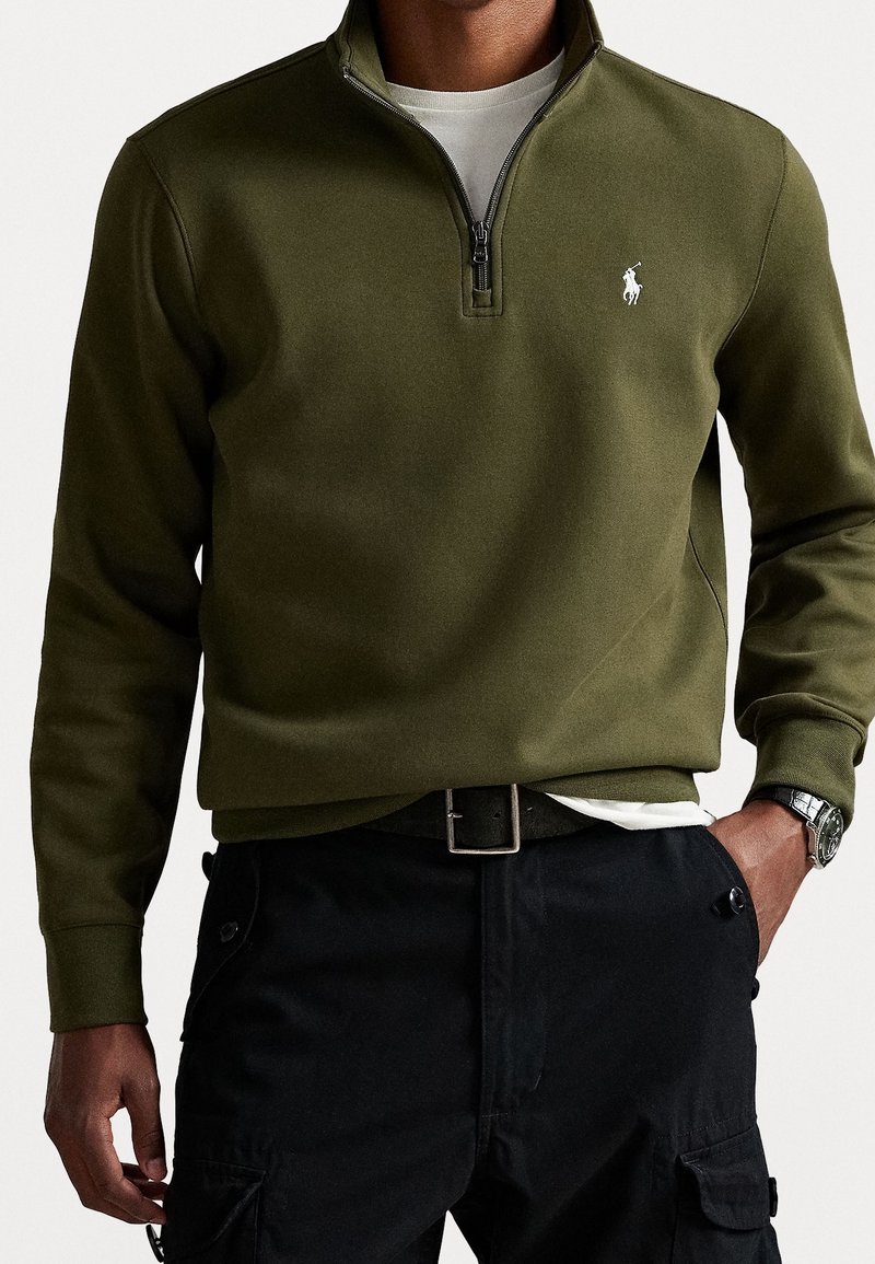 Homme portant un sweat-shirt kaki à fermeture éclair quart de zip avec logo blanc, pantalon noir avec poches et montre-bracelet argentée, main dans la poche.