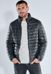 Schwarze gesteppte Steppjacke aus glänzendem Material mit Stehkragen, Frontreißverschluss und Seitentaschen. Getragen über einem hellgrauen Hemd.
