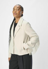Blazer color marfil con cuello clásico, cierre de botones y detalles de flecos en los puños. Textura suave y silueta estructurada.