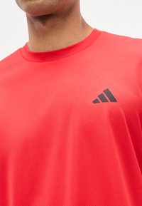 Personne portant un t-shirt de sport rouge vif avec un logo Adidas noir sur le côté gauche de la poitrine, gros plan montrant l'encolure et le haut du torse.