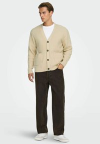 Beige Rippenstrickjacke mit V-Ausschnitt, drei vorderen Knöpfen und zwei vorderen Taschen, kombiniert mit dunklen Hosen und weißen Sneakern.