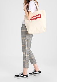 Beżowa torba tote z płótna z czerwonym logo "Levi's", noszona na ramieniu. Model w kraciastych spodniach i czarnych sneakersach stoi na szarym tle.