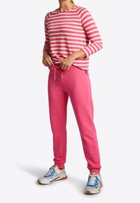 Pantalons de survêtement roses avec taille à cordon, associés à un t-shirt à manches longues rose et blanc à rayures. Le mannequin porte des baskets blanches avec des accents multicolores.