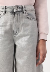 Mittelteil einer Person, die hoch taillierte hellgraue Jeans und ein blassrosa Langarmshirt trägt, Hand entspannt an der Seite.