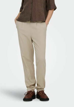 Persona vestida con pantalones beige, camisa marrón con botones y zapatos marrones de cordones, de pie con las manos en los bolsillos frente a un fondo liso.