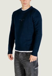 Felpa a maglia blu navy con collo e polsini a coste, caratterizzata da un piccolo logo ricamato sul petto, abbinata a jeans scuri.