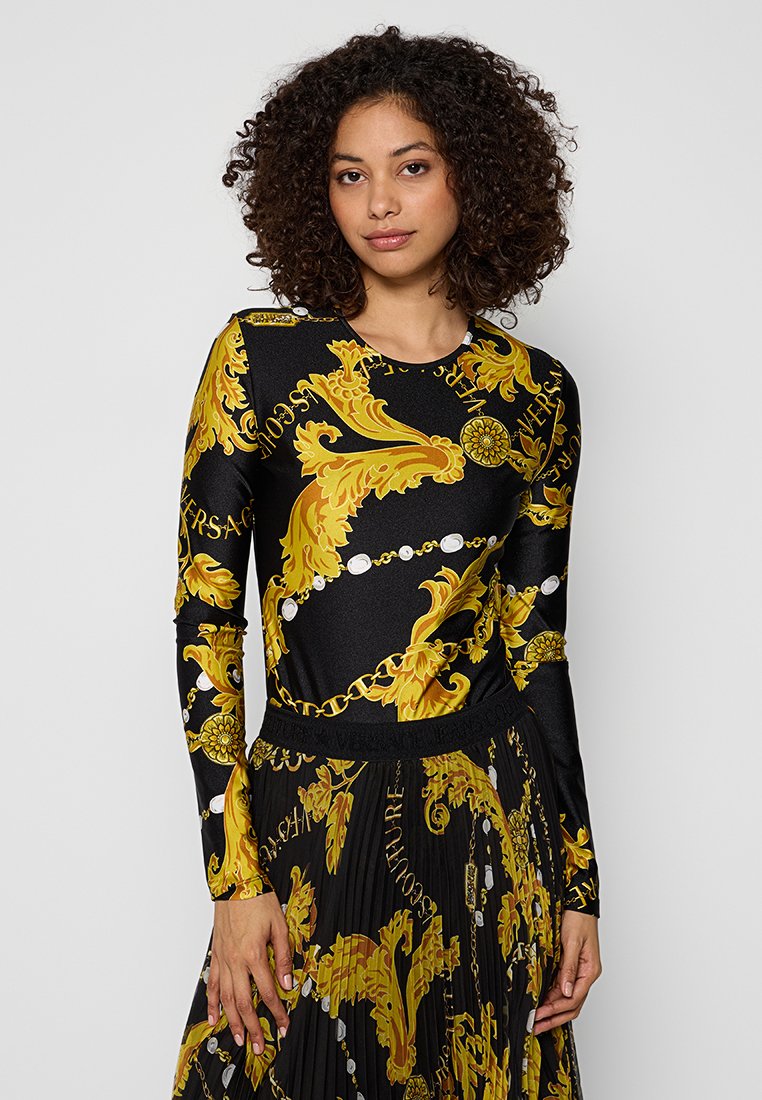 Femme aux cheveux bouclés portant un haut noir à manches longues et une jupe plissée ornée d'un motif baroque en or et de chaînes.