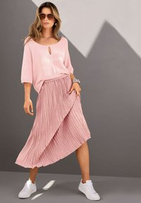 Helles rosa gestricktes Oberteil mit Schlüsselloch-Detail, kombiniert mit einem plissierten rosa Rock. Weiße Turnschuhe vervollständigen den Look und zeichnen sich durch glatte Texturen aus.