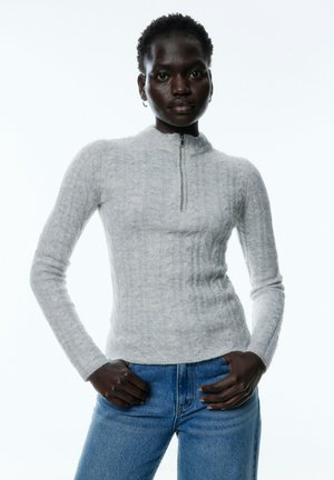 Grijze kabelgebreide trui met een hoge hals en rits. Aansluitend ontwerp met lange mouwen, gedragen over blauwe denim jeans.