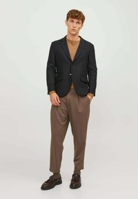 Blazer scuro texturizzato con due tasche frontali, abbinato a un maglione marrone e pantaloni marroni larghi. Gli accessori includono scarpe marroni.