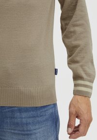 Beige kötött pulóver bordás szegéllyel és mandzsettával; krémszínű és világosbarna csíkos díszítés. Puha tapintás, testhezálló fazon, kék farmernadrággal párosítva.